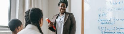 Teacher smiling at students, standing beside whiteboard with questions written on it