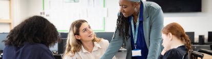 Teacher leaning over desk pointing to sheet. Student looking up at teacher.